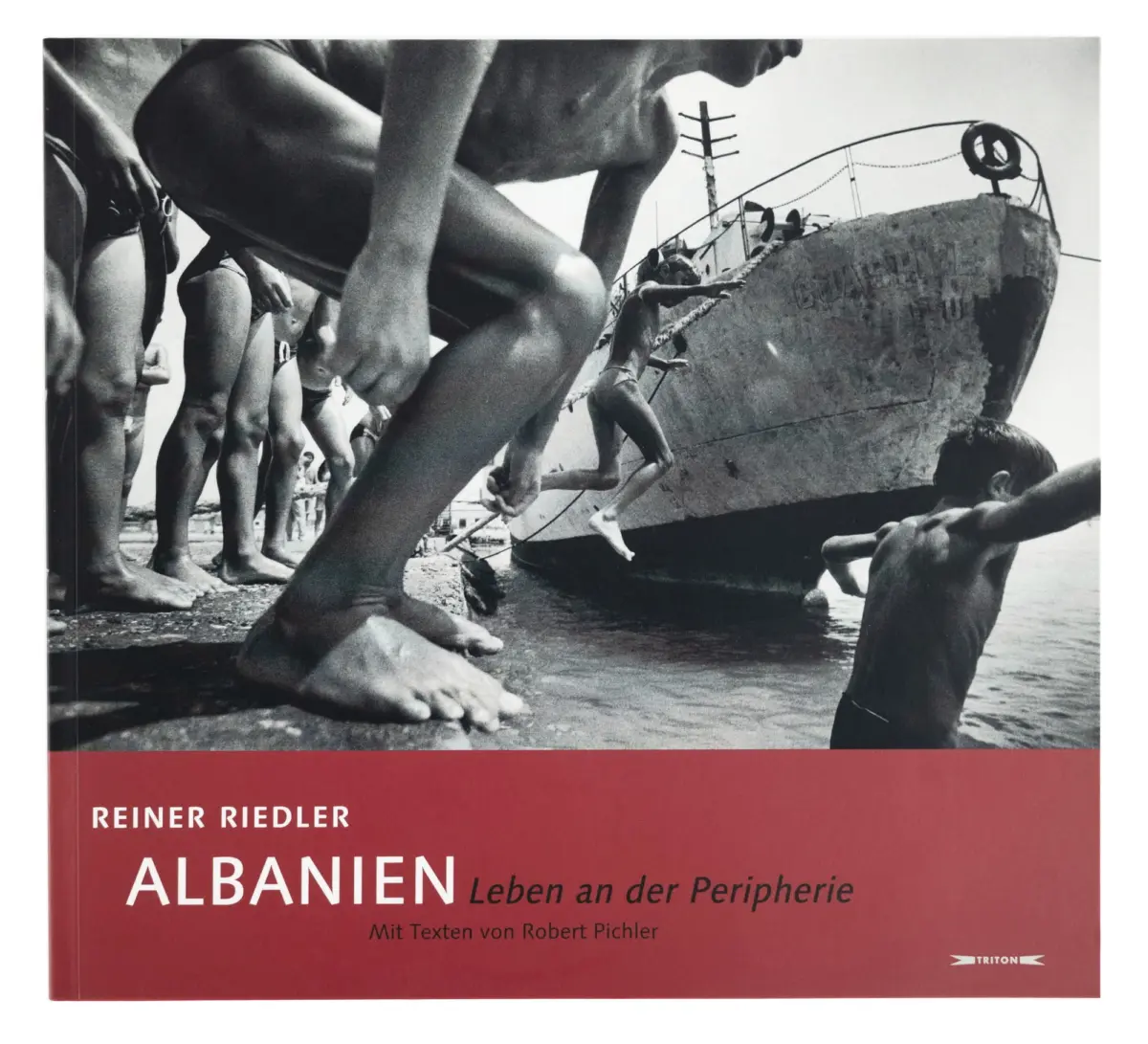 Black-and-white photograph of swimmers lined along a beach, scrambling onto a large rusted ship hull at the water's edge.