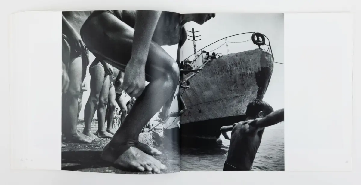 Black-and-white beach scene: a line of barefoot beachgoers reach toward a weathered, partially submerged boat at the water's edge.