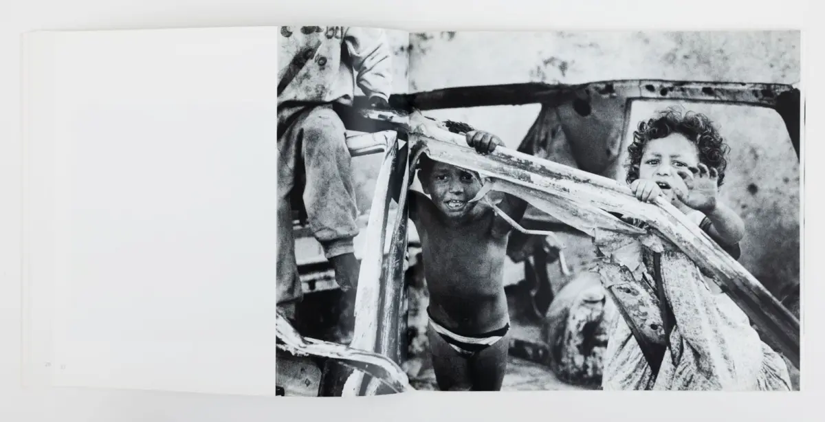 Two children grip a torn metal frame of a wrecked vehicle outdoors; black-and-white photograph.