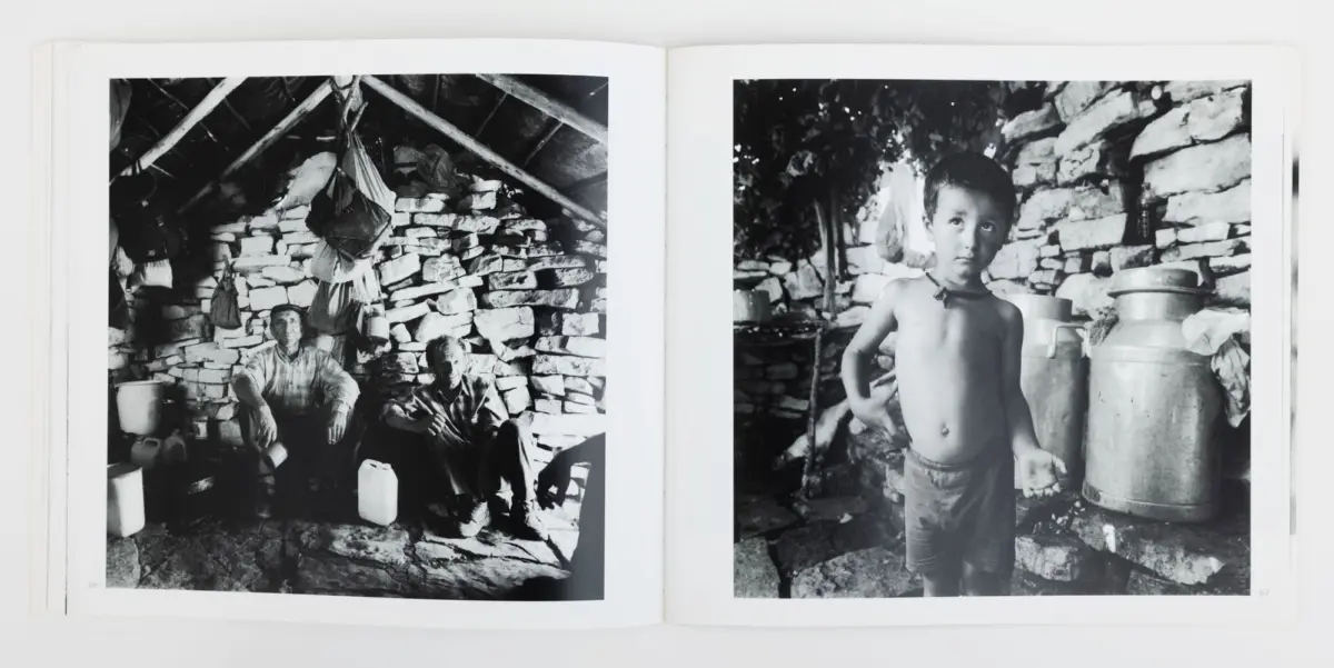 Two men sit inside a stone-walled hut with hanging bags; a shirtless boy stands beside a large metal milk can near a stone wall.