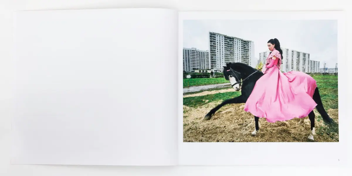 Person in a bright pink gown rides a black horse across a dirt field with high-rise apartment buildings in the background.
