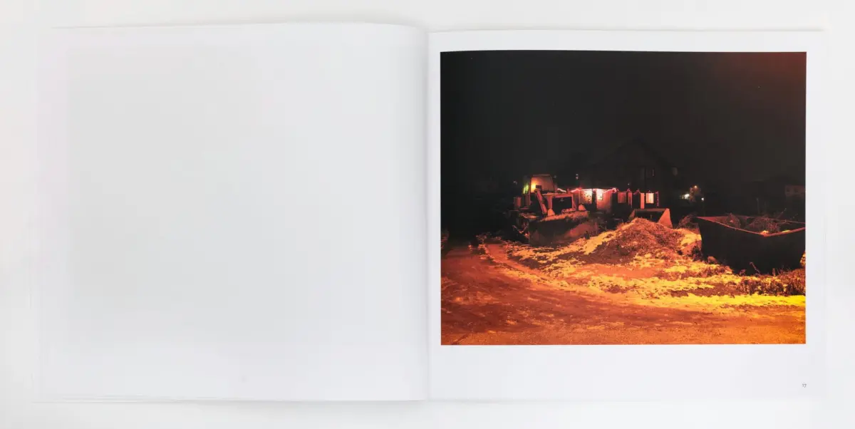Nighttime construction site with an excavator, snow-covered ground, and piles of earth; distant houses glow with orange lights.