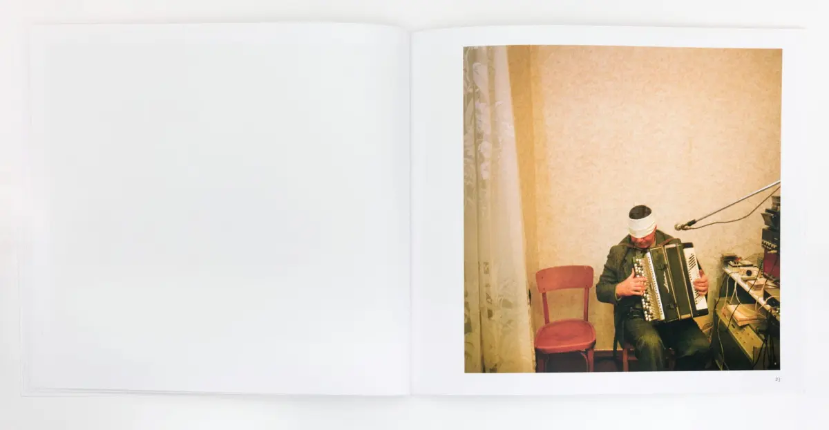Man with head bandage plays accordion in a small room with beige wallpaper and red chairs.