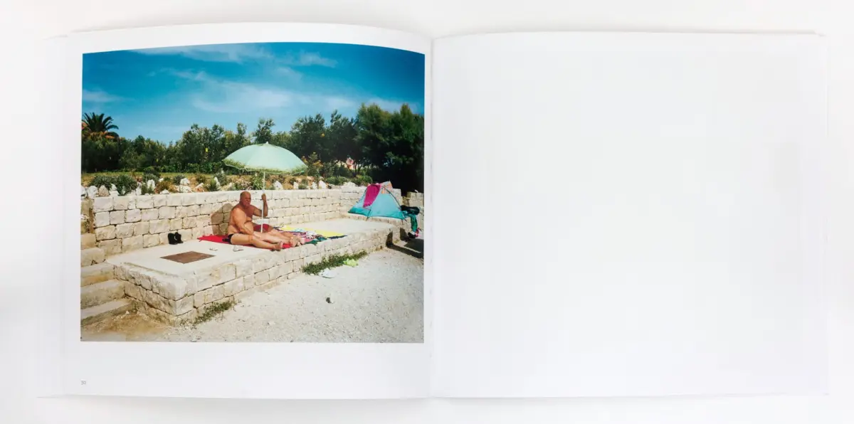 Adult sunbather lying on towel on a stone garden terrace under a green umbrella; a turquoise tent and towels nearby.