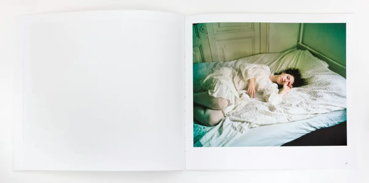 Person lying on bed in white nightgown, resting on white bedding in a green-walled bedroom.