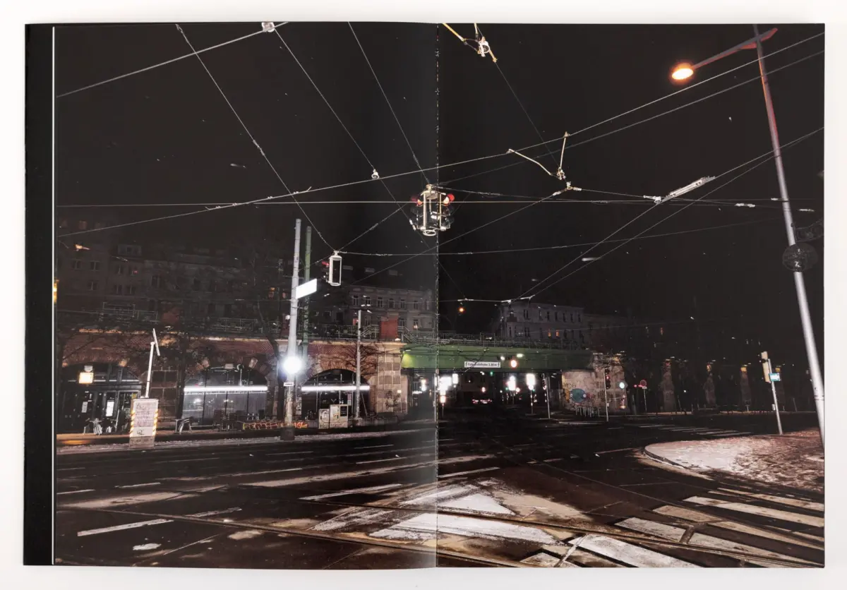 Nighttime urban intersection with crisscrossing tram cables, a traffic light, and brick buildings along a wet street.