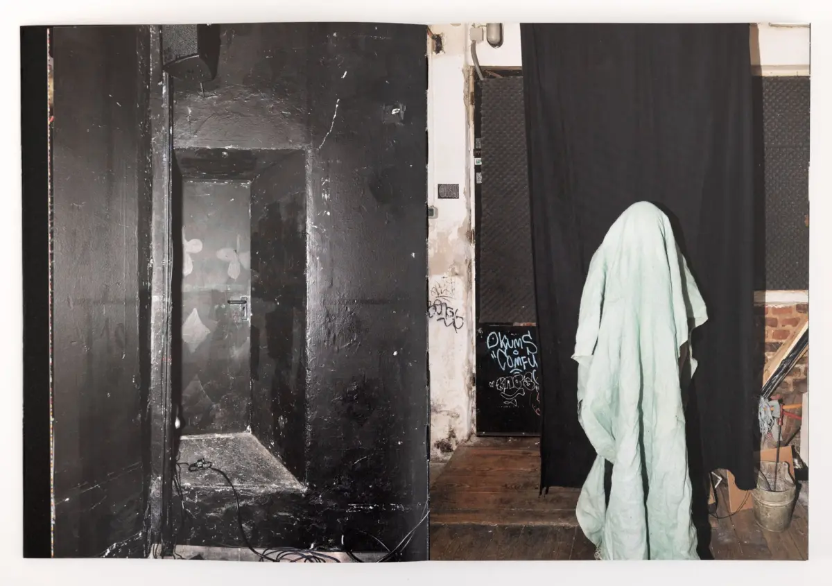 Person draped in a pale green sheet stands beside a black curtain in a gritty backstage area with exposed walls and tangled cables.