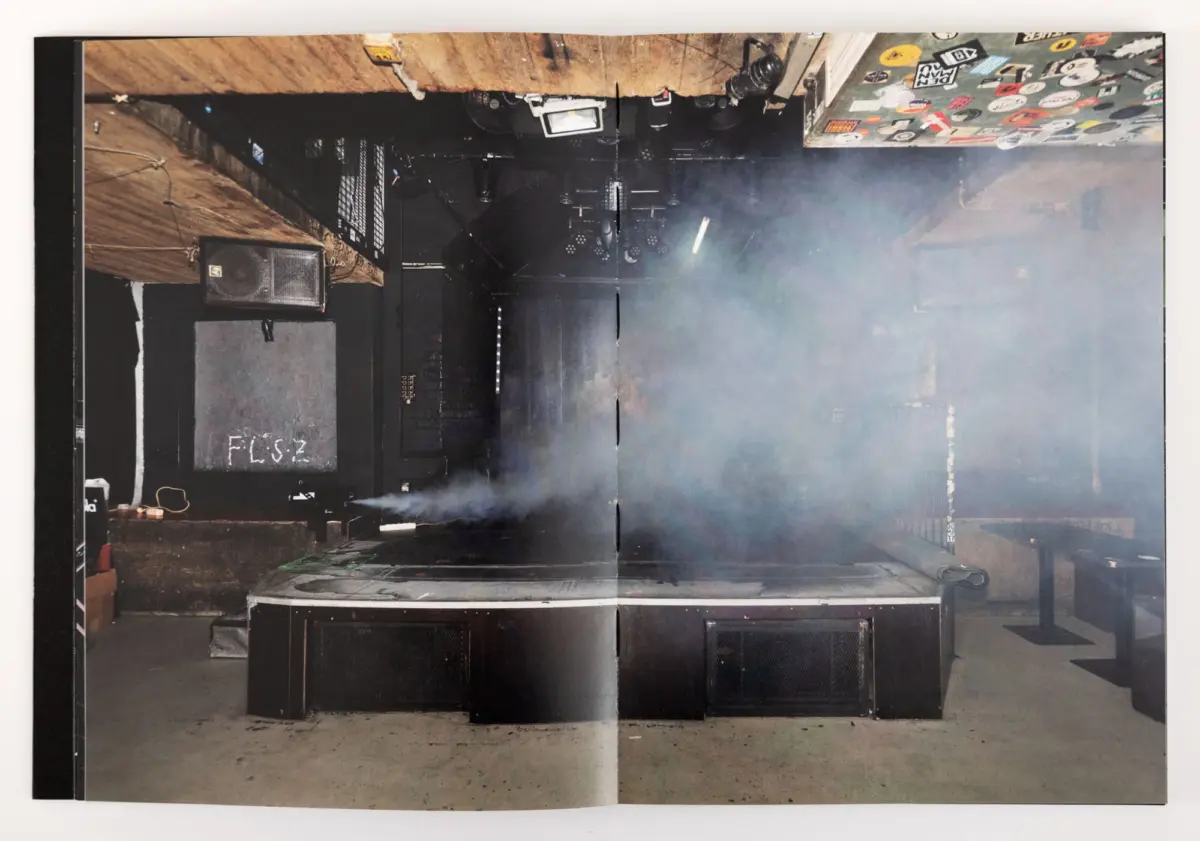 Concert stage shrouded in smoke with large black speaker cabinets, lighting rigs, and scattered equipment.