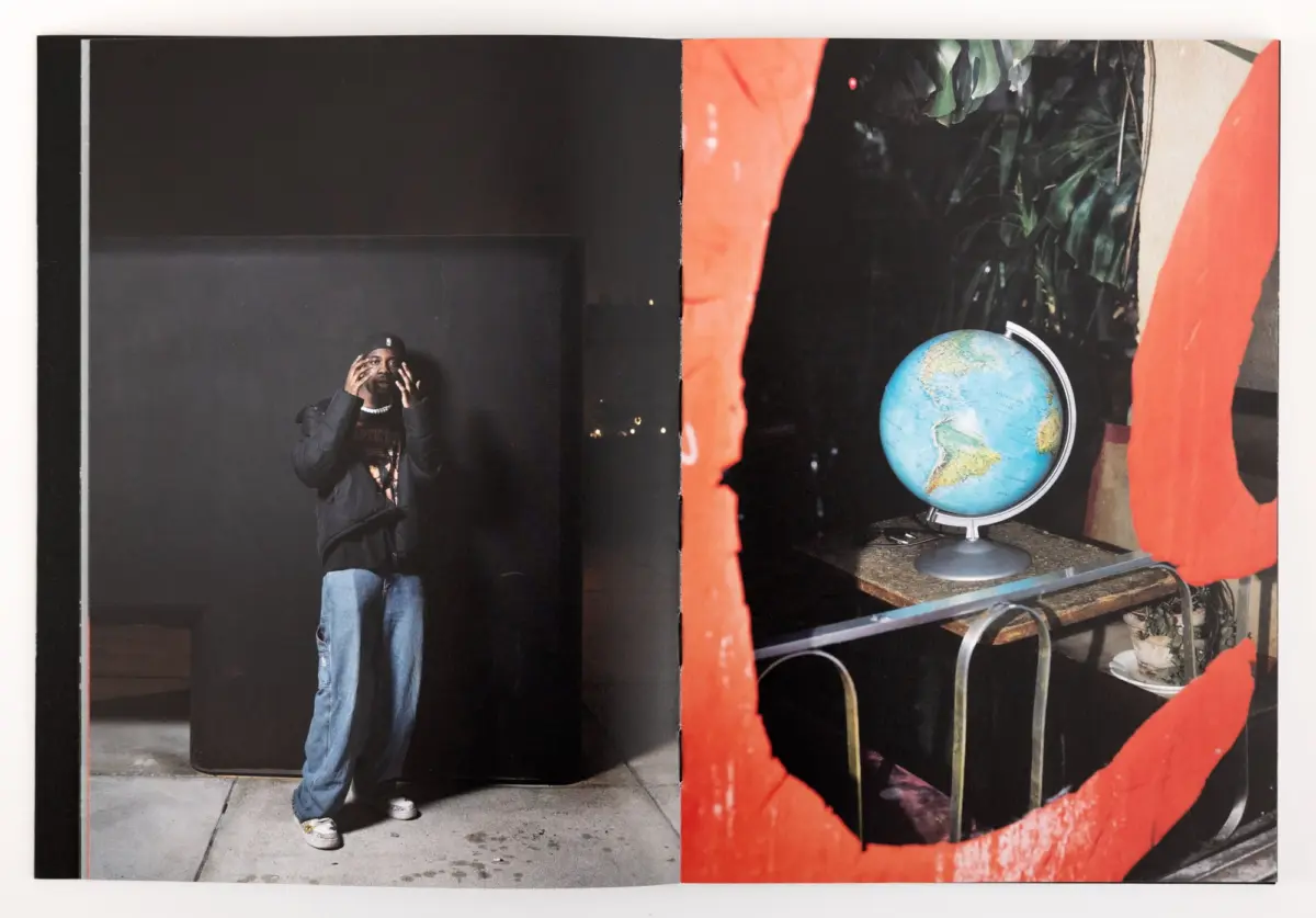 Person wearing a cap and dark jacket stands in a black-walled space, hands raised near the face; to the right, a globe on a desk beside a red mural.