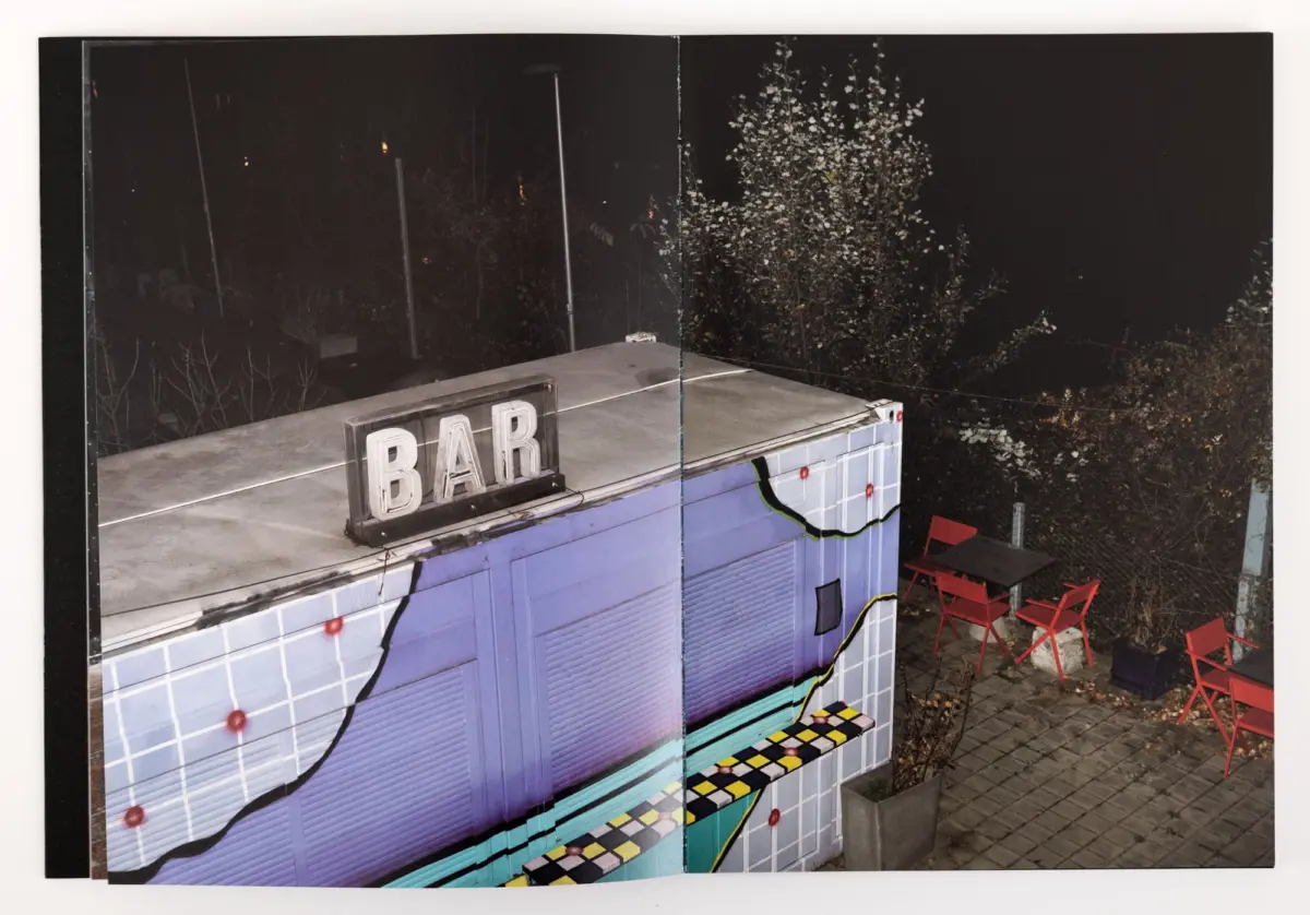 Night scene of a pastel-blue bar counter with a BAR sign; red outdoor chairs nearby and trees in the dark background.