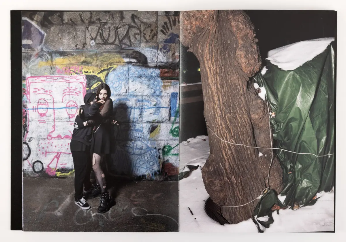 Young person in black points at camera beside graffiti-covered wall; snow on ground, large tree trunk, and green tarp.