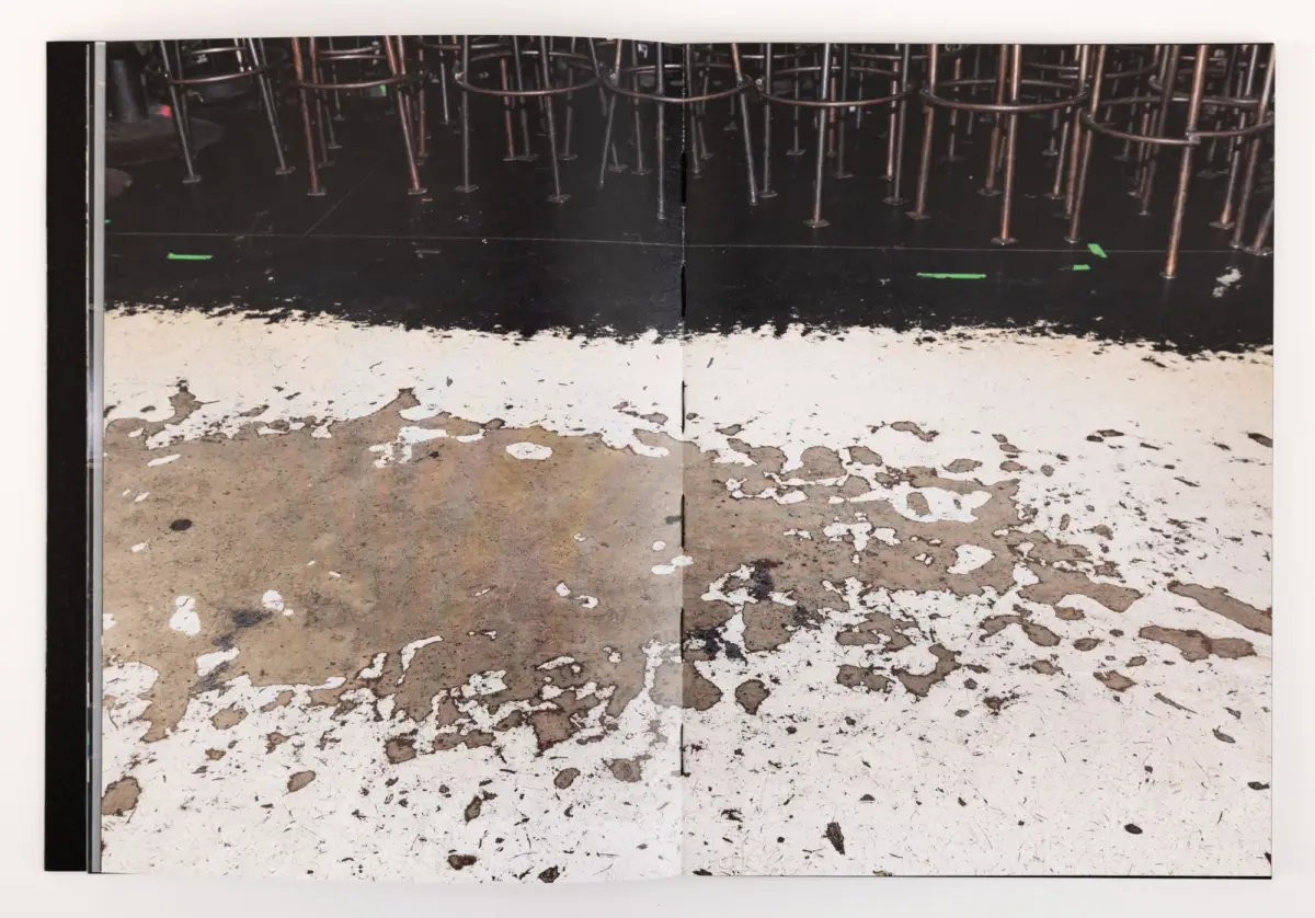 Underside view of numerous metal chair legs on a dark stage, with green tape marks and a peeling, speckled floor.
