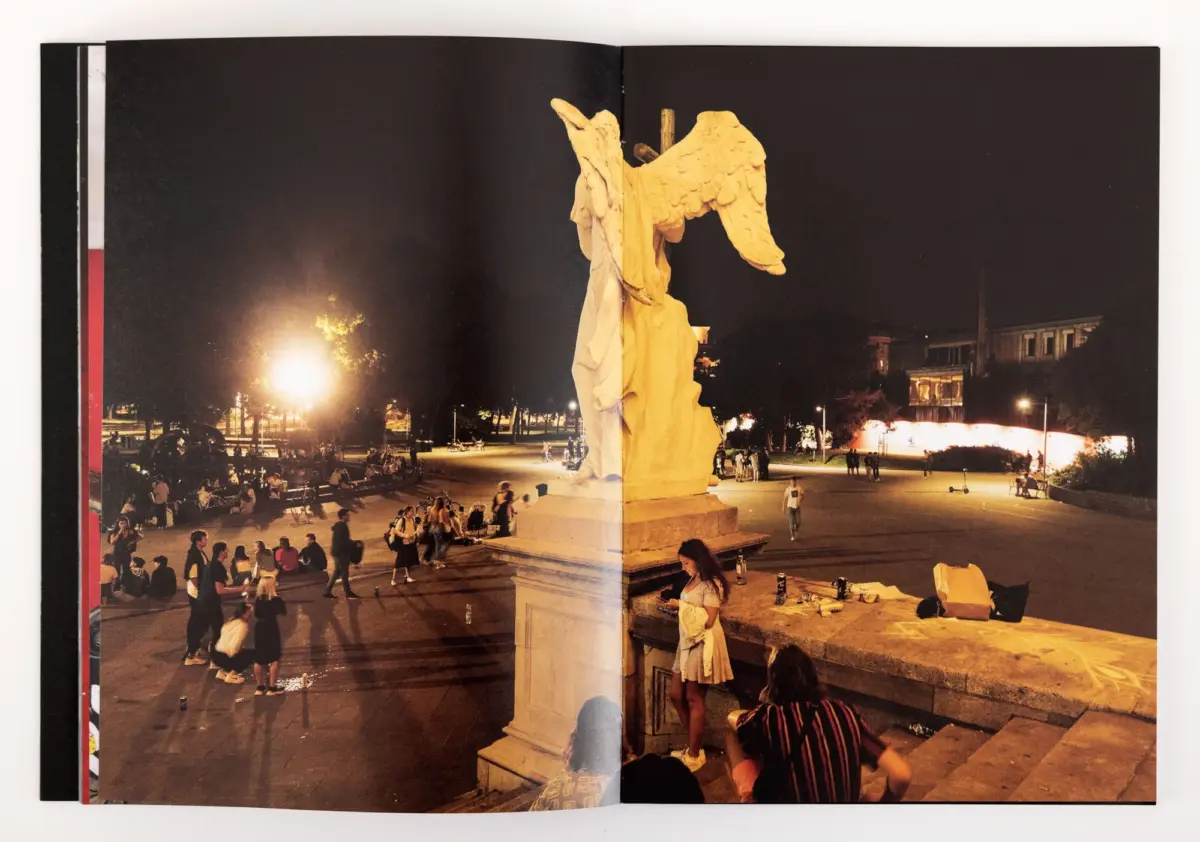 Nighttime city plaza with a tall winged angel statue on a pedestal; people gather and stroll as lights illuminate surrounding buildings.
