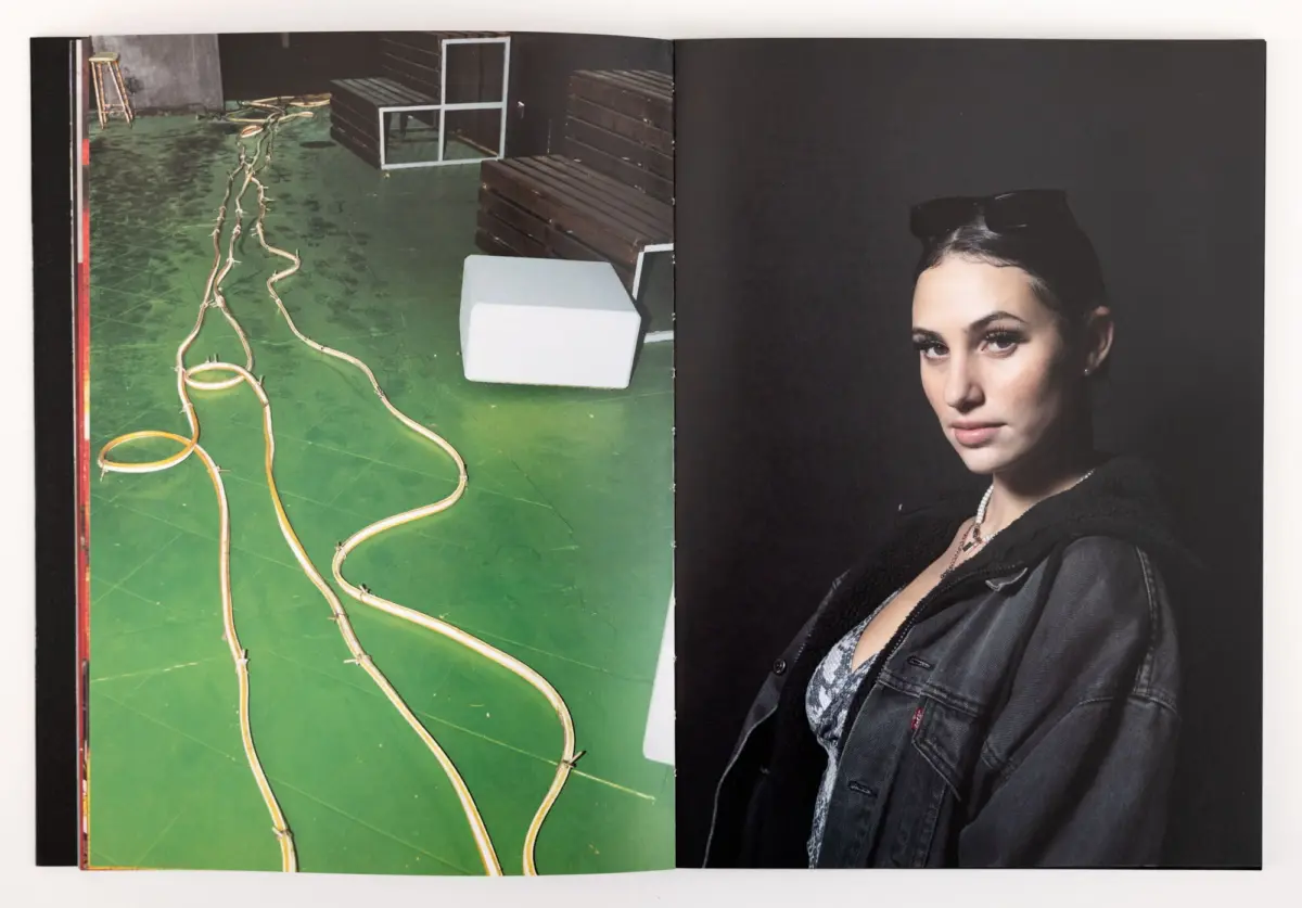 Studio portrait: person in denim jacket with sunglasses perched on head; left side shows green floor, tangled cords, white block, and stacked benches.