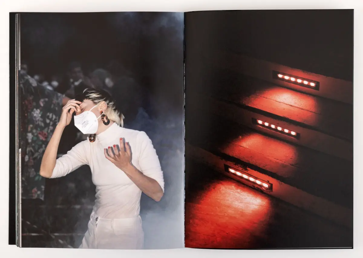 Woman performer in white outfit and face mask on a smoky stage; red-lit stairs on the opposite page.