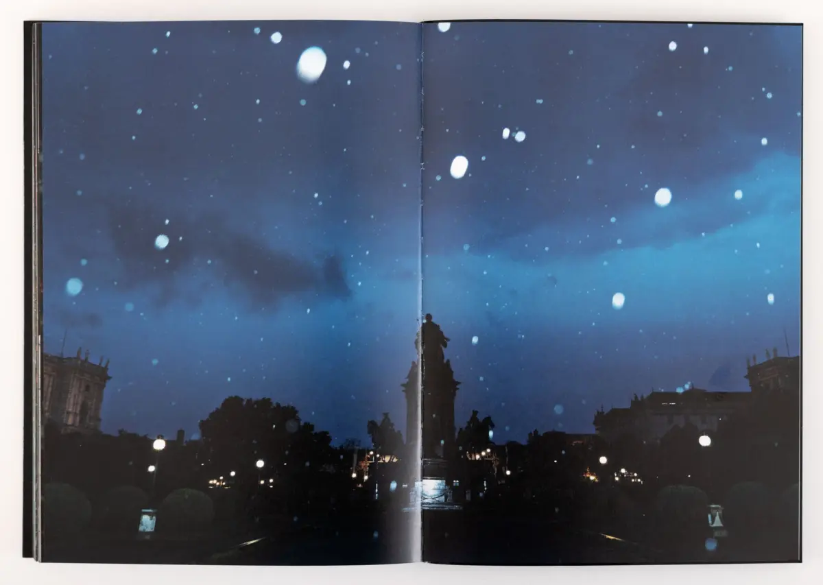 Nighttime city square with a tall obelisk monument, silhouettes of people, and falling snow.