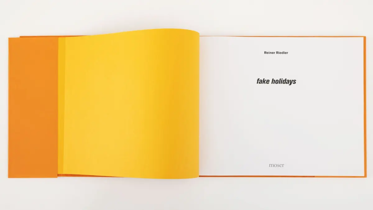 Open book with orange cover and yellow pages; right page displays bold title fake holidays by Reiner Riedler.