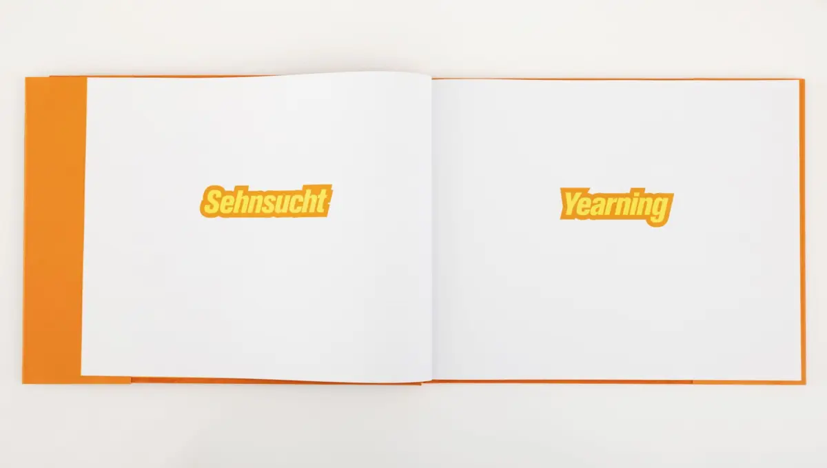 Open orange hardcover book with two white pages; left page reads Sehnsucht, right page reads Yearning.