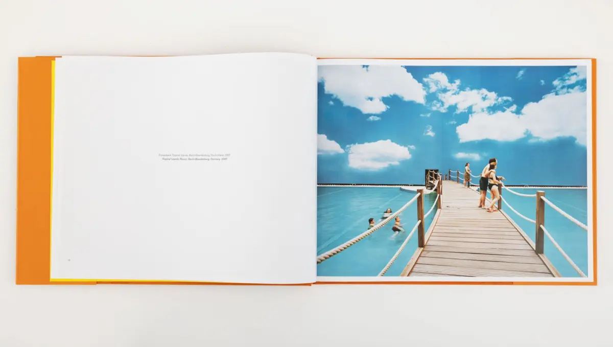 Open book page showing a wooden pier over turquoise water with people walking and swimming beneath a bright blue sky.