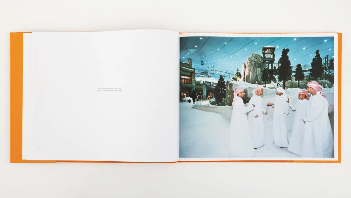 Open book; right page shows men in white kanduras and red ghutras greeting each other in an indoor snowy ski area with rocks and a lookout tower.