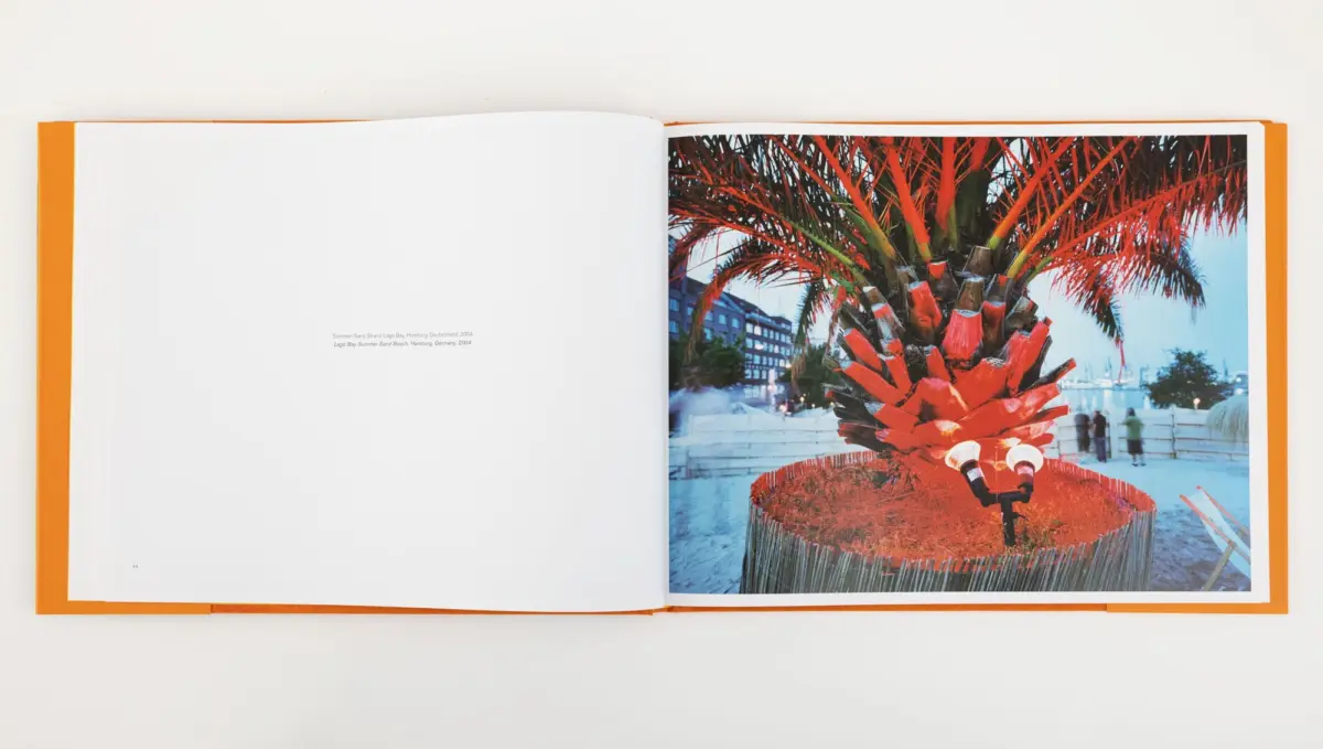 Open book page showing a red-painted palm tree inside a circular fence at a waterfront plaza, with buildings and people in the background.