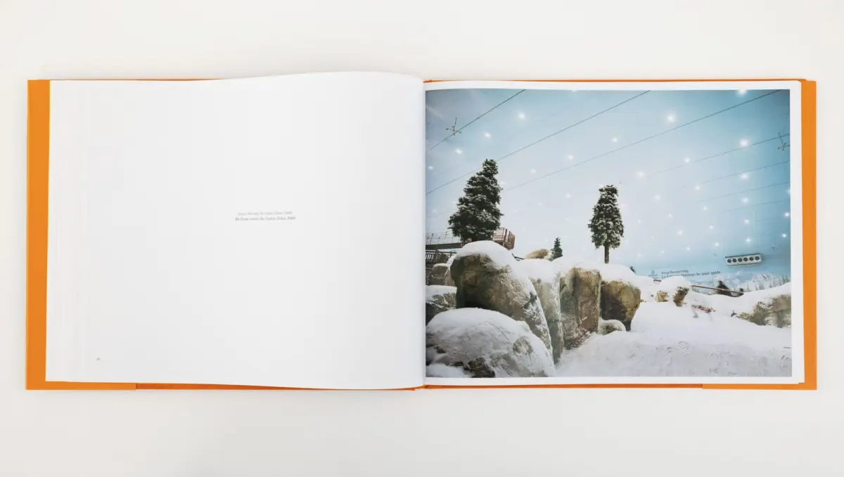 Open orange-bound book; right page shows an indoor snowy landscape with rocks, evergreen trees, and overhead ceiling lights.