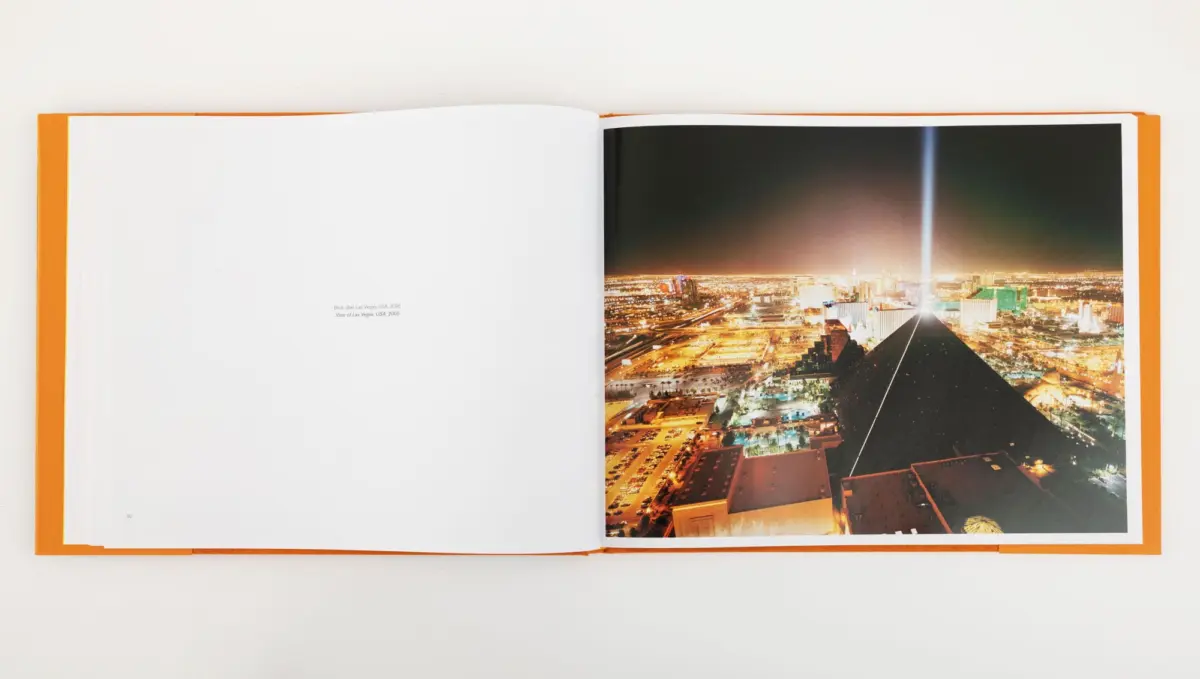 Open orange-bound book on white surface; right page shows a nighttime cityscape with a bright vertical beam of light.