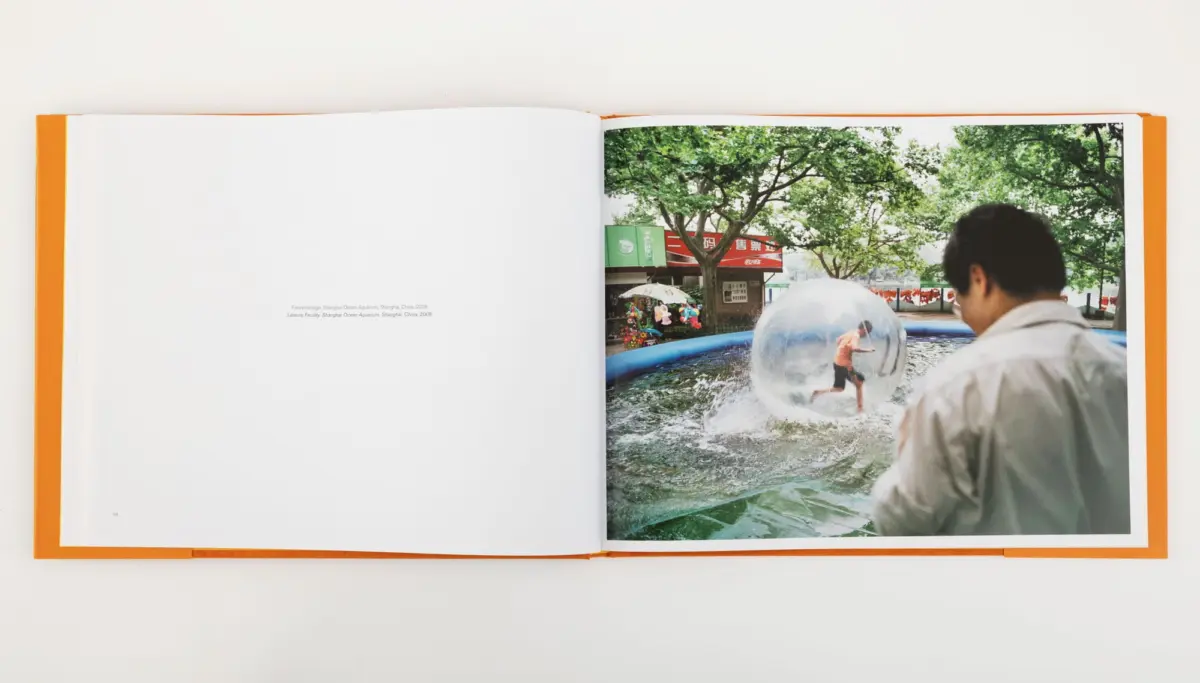 Child inside a giant transparent inflatable ball rolls in a shallow pool under trees; nearby adult man observes.
