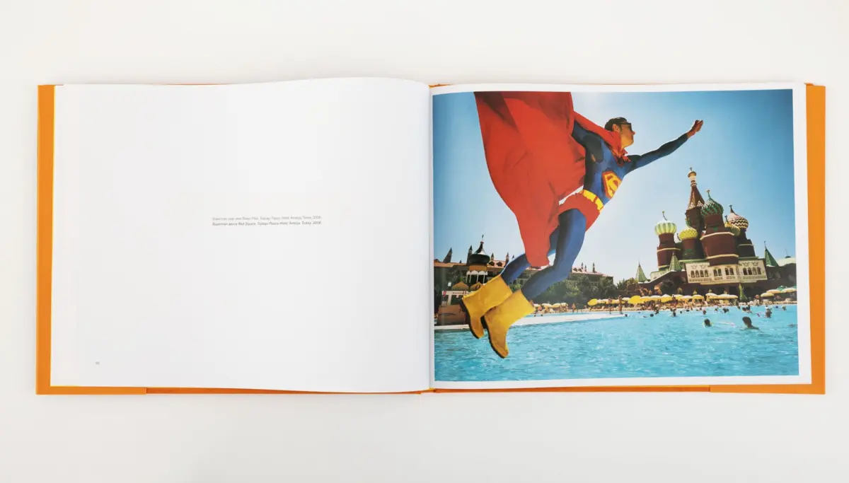 Superman in blue suit and red cape leaps over a swimming pool at a Moscow water park, with Saint Basil's Cathedral in the background.