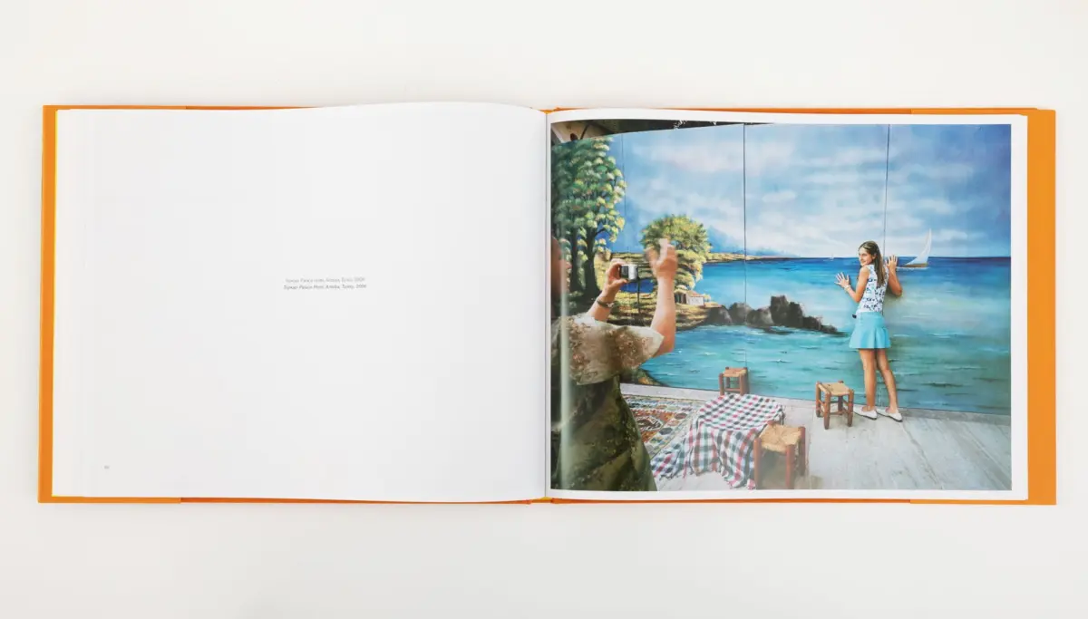 Open book with orange cover; mural shows a girl in a blue skirt by the sea, as a photographer in the foreground takes a photo.