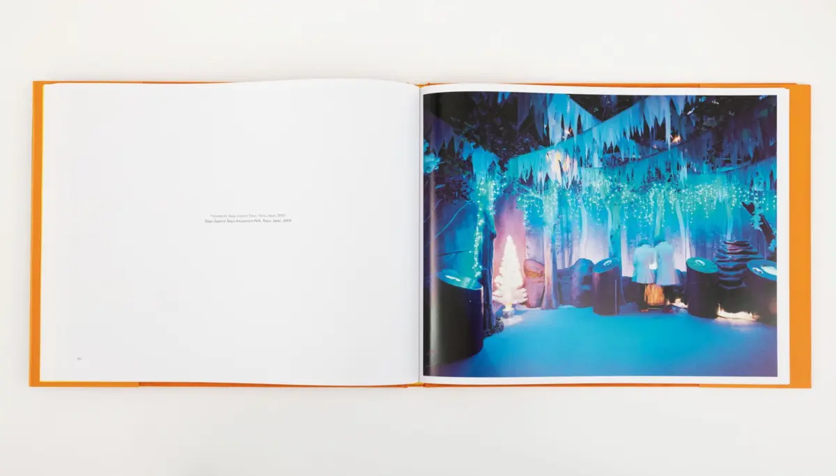 Open photo book with orange cover; right page shows blue-lit forest party scene with pennant garlands, string lights, and silhouettes of people.