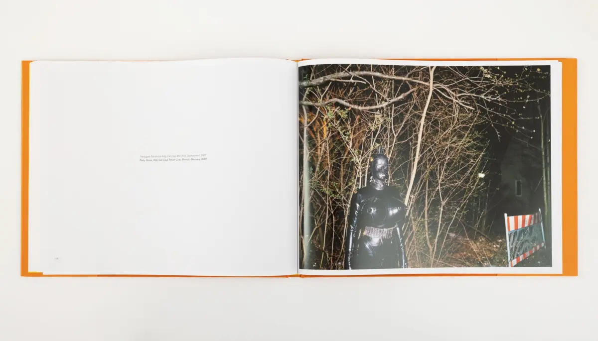Person in black latex suit and mask stands among bare branches at night, with a striped orange barrier nearby.