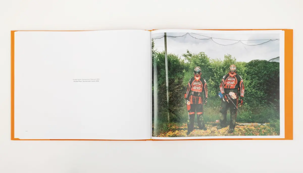 Open book page showing two paintball players in red and black gear with masks, standing outdoors among greenery.
