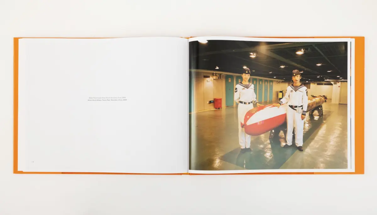 Two sailors in white naval uniforms pose with a large red-and-white torpedo in a naval maintenance hangar.