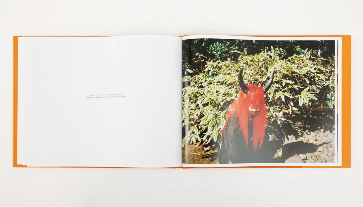 Person wearing a red wig and black horned headpiece, posing outdoors among green bushes.