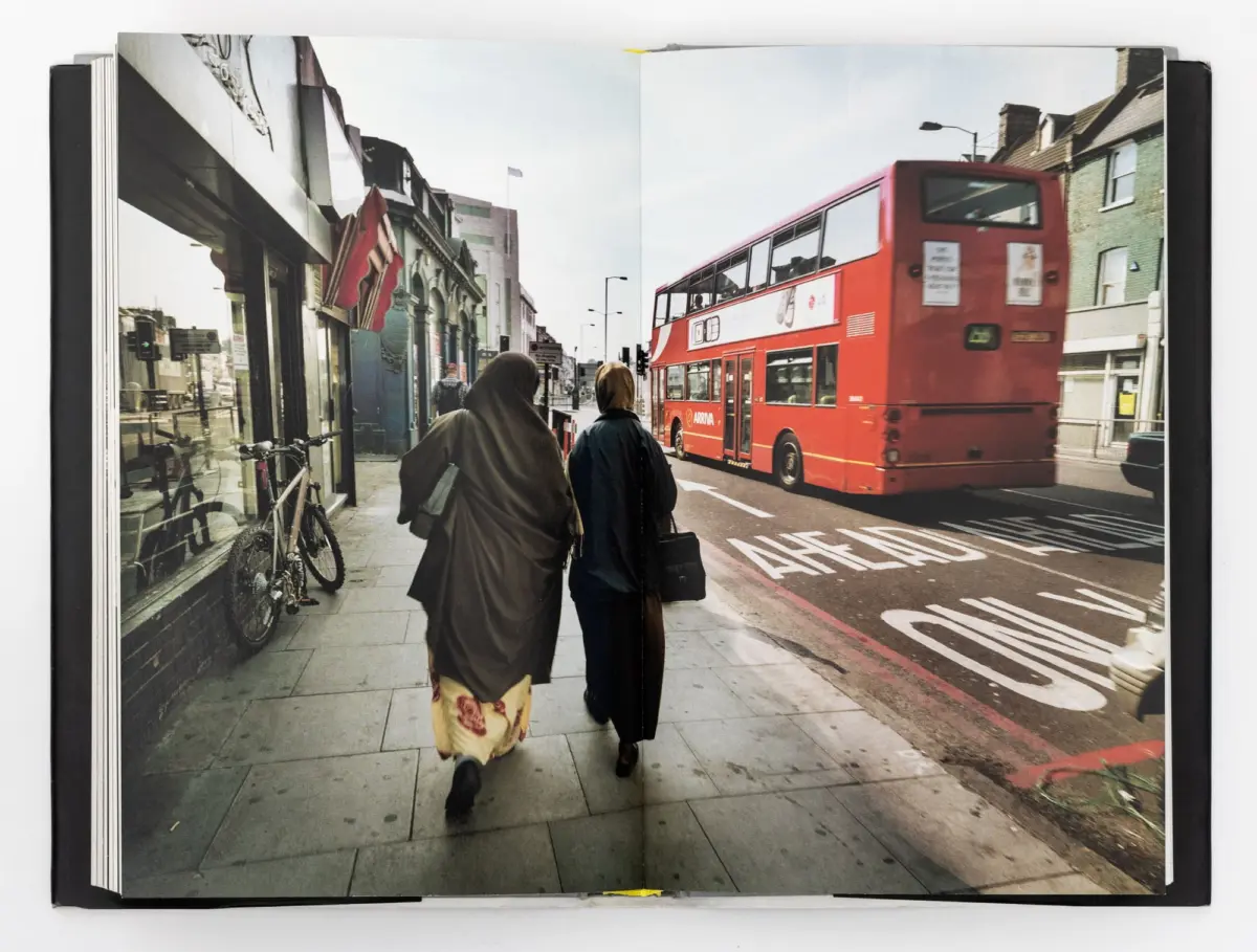 Two women wearing hijabs walk away from the camera along a city sidewalk, beside a red double-decker bus and row of shops.