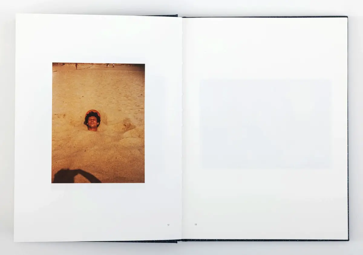 Left-page photo of a man partially buried in beach sand, head and shoulders exposed; open book with blank right page.