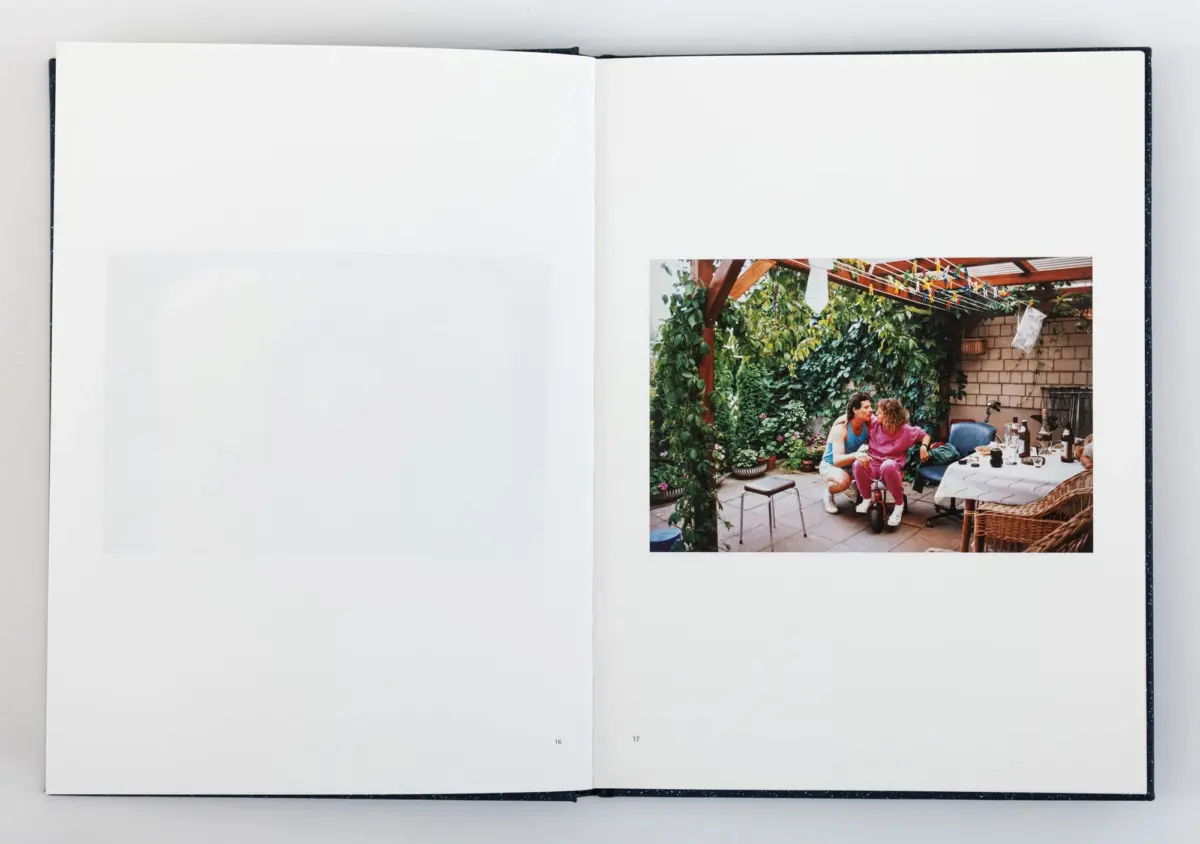 Two adults sit at a patio table in a leafy backyard garden under a wooden pergola, with dishes, drinks, and a clothesline overhead.