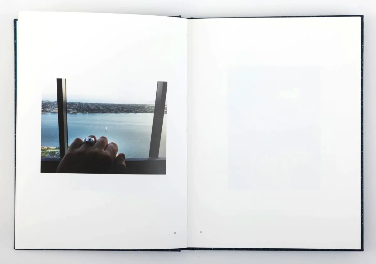 Hand holding a camera at a balcony railing, framing a harbor view with water, a sailboat, and distant city skyline.