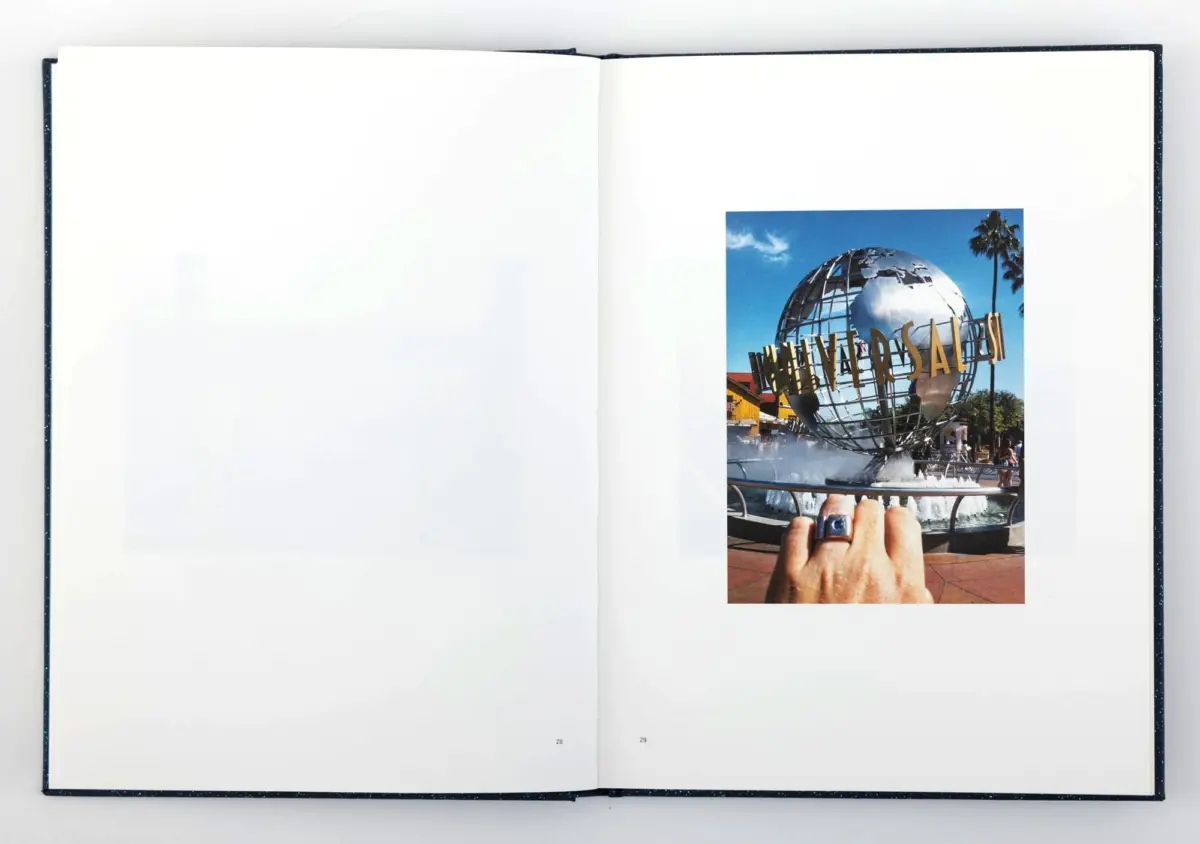 Open book showing a right-page photo of a chrome globe sculpture, palm trees, fountain, with a hand wearing a blue ring in the foreground.