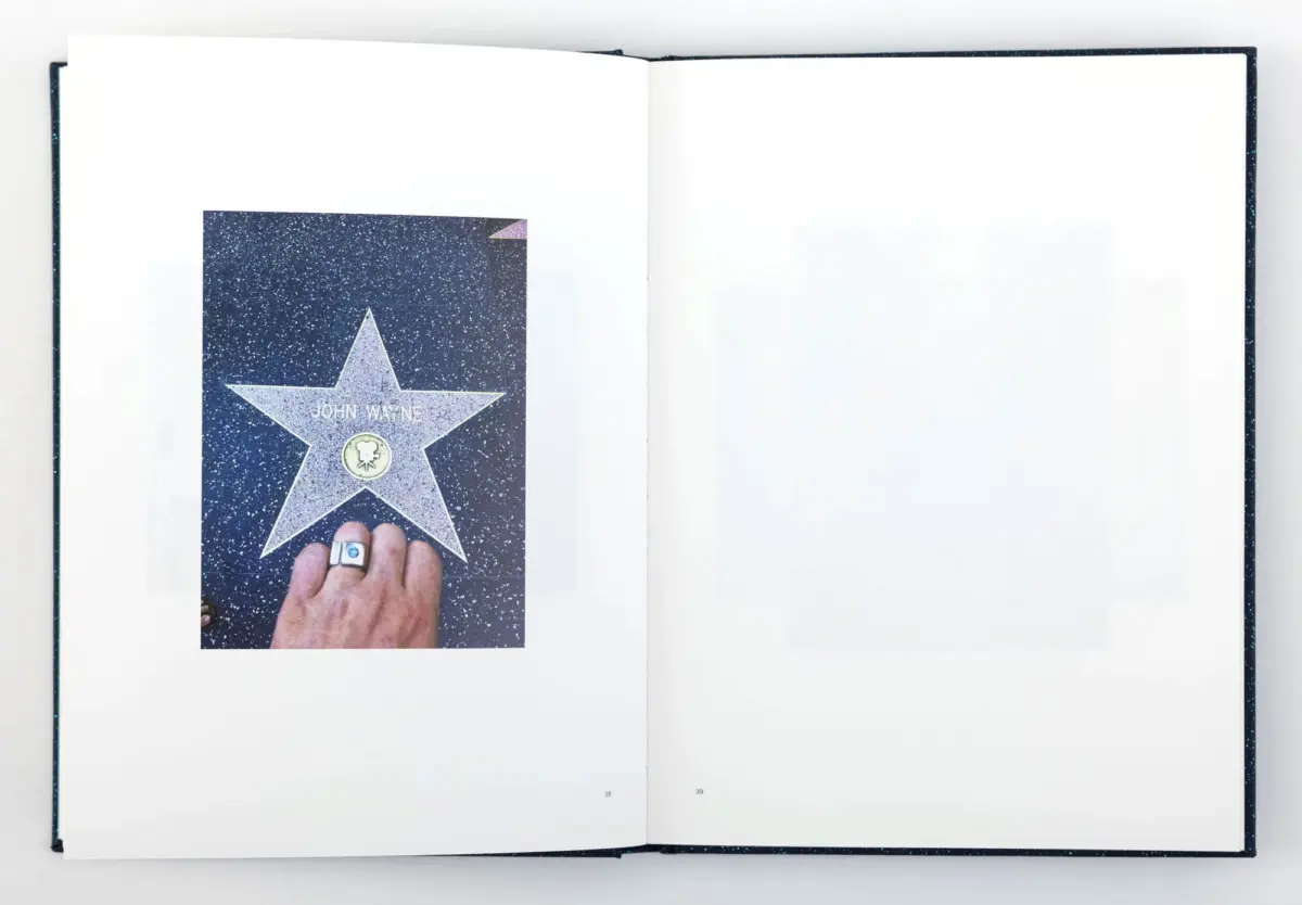 Hand with ring holds a John Wayne Walk of Fame star card on the left page of an open book.