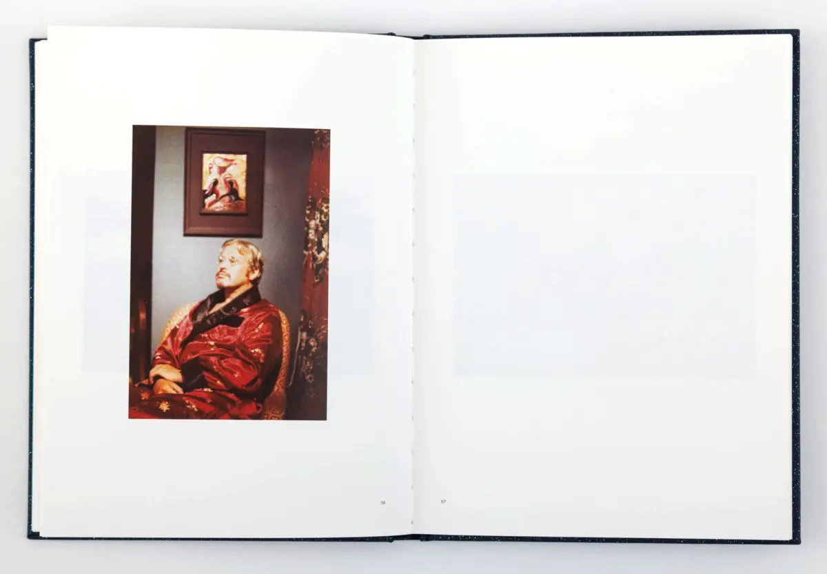 Photograph of a person in a red robe seated indoors, on a page of an open book, with a framed painting of birds on the wall behind.