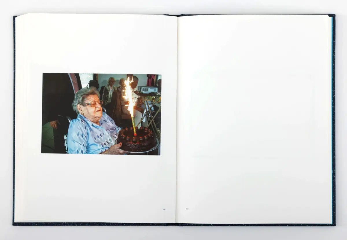 Older person in a blue patterned shirt holds a chocolate birthday cake with a tall lit candle; party guests in the background.