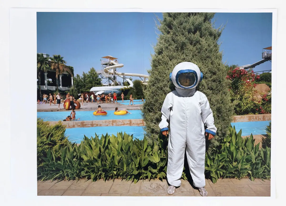 Person in white spacesuit standing in front of a water park with slides, pool, and greenery in the background.