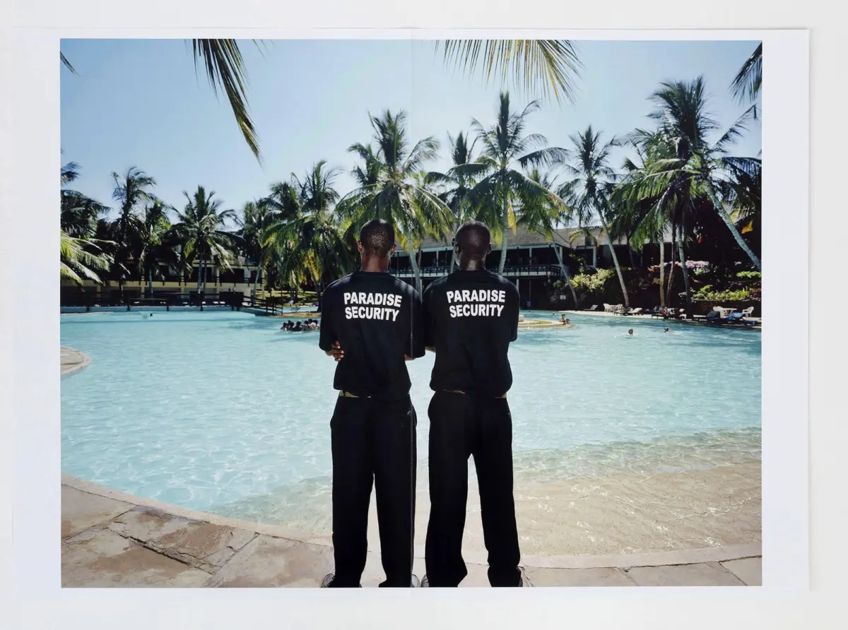 Two security guards in black shirts with 'Paradise Security' stand with backs to the camera by a tropical resort pool with palm trees.