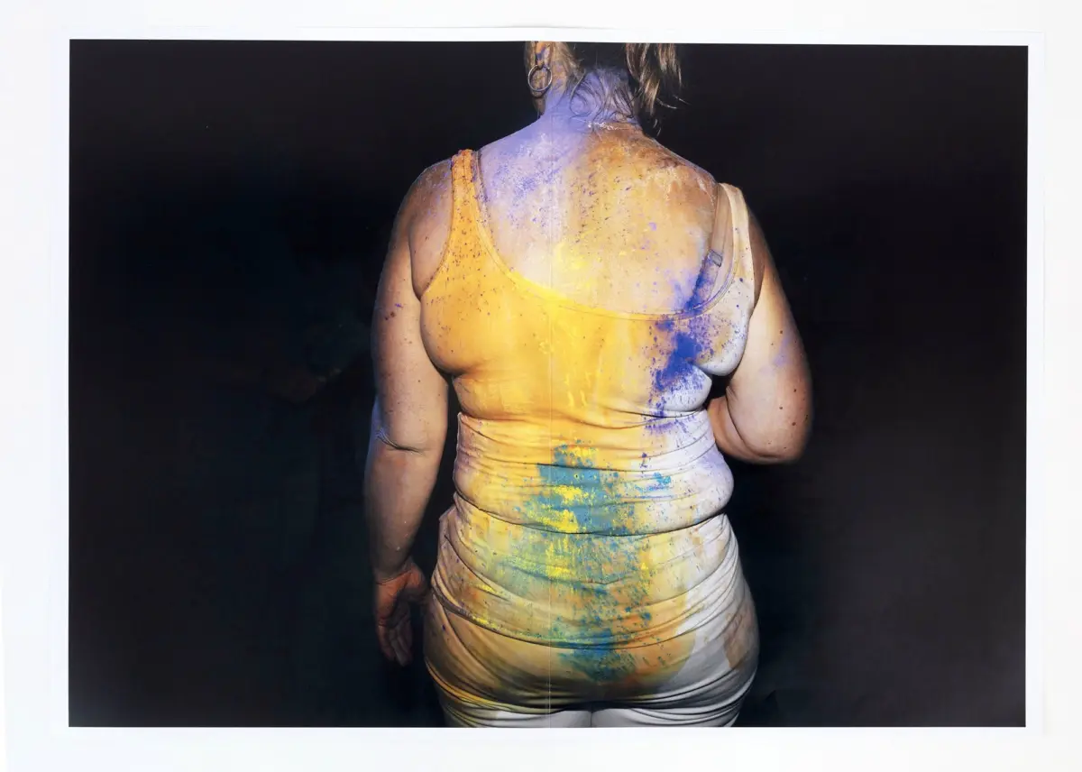 Adult person seen from behind in a yellow tank top splattered with paint, standing against a dark backdrop.