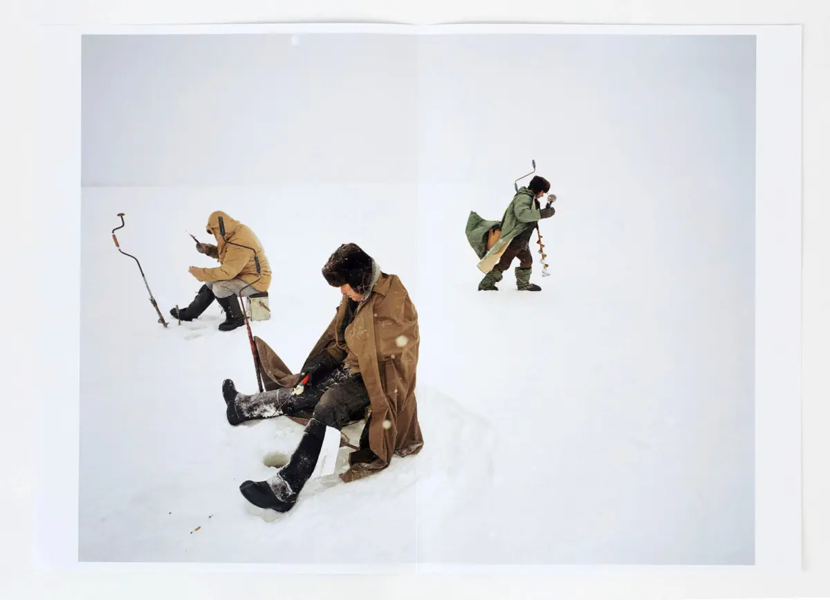 Three people in heavy winter coats on a snow-covered landscape; one sits, another kneels with a walking stick, the third stands with an ice auger.