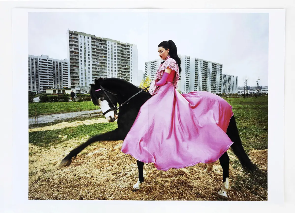 Person in a pink ball gown rides a black horse across a dirt field with high-rise apartment buildings in the background.