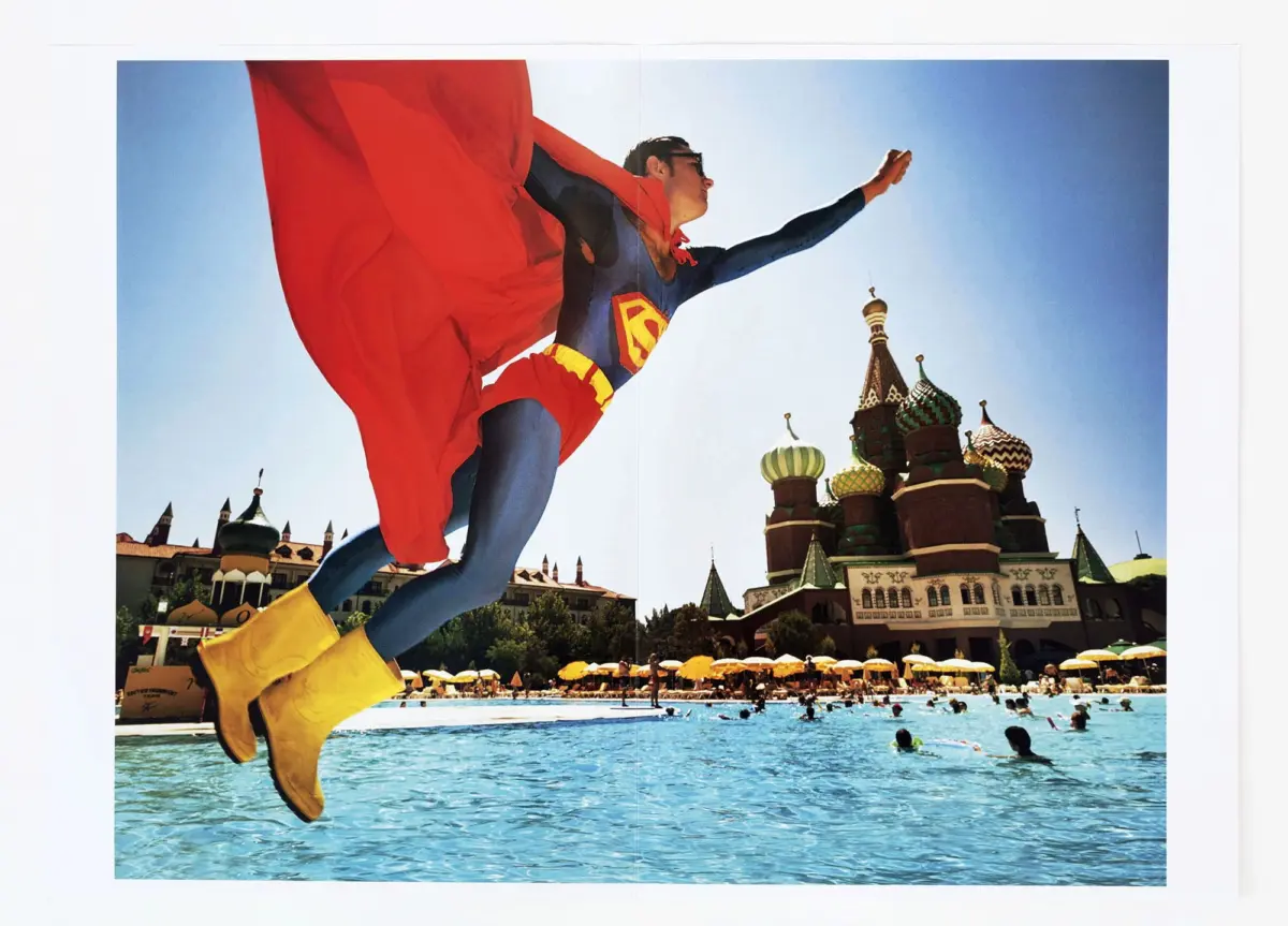 Costumed superhero in blue suit and red cape flies over a crowded pool, with onion-domed cathedral in the background.