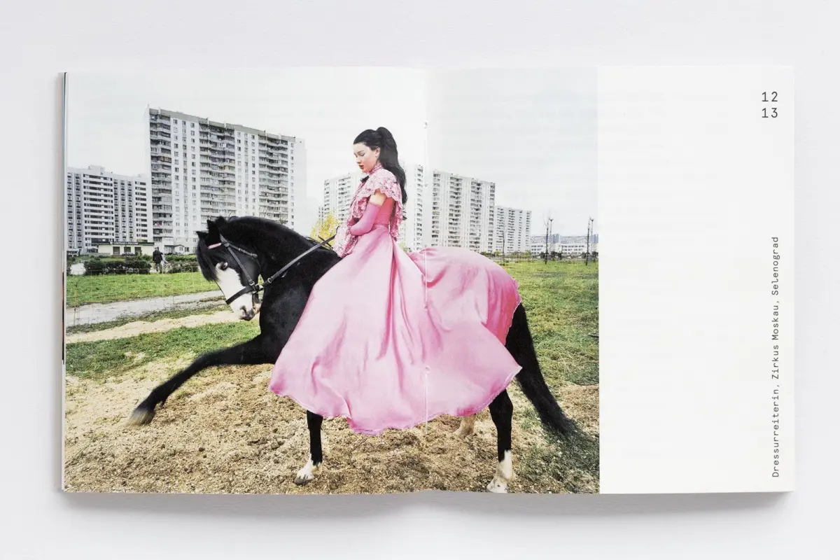 Person in a pink ball gown rides a black horse across a dirt field with high-rise apartment buildings in the background.